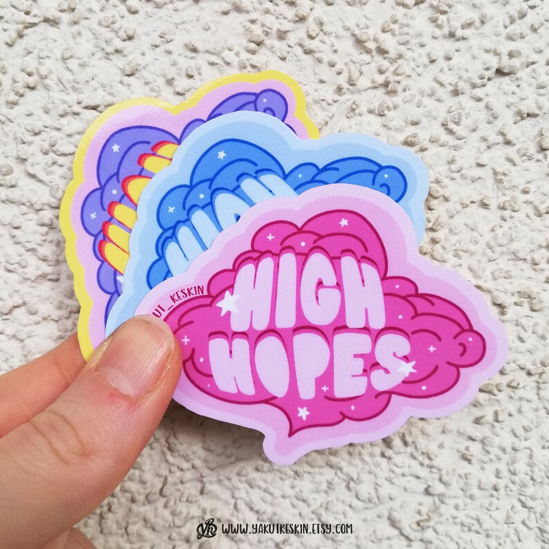 Puede incluir: Tres pegatinas coloridas con un dise&ntilde;o de nube. La pegatina superior es amarilla con una nube morada y el texto "Stay Wild". La pegatina del medio es azul con una nube azul y el texto "Dream Big". La pegatina inferior es rosa con una nube rosa y el texto "High Hopes".