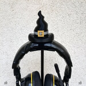 May include: Black headphones with a black witch hat on the stand. The hat has a silver band and a gold letter "B" on the front.