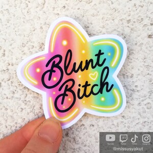 May include: A rainbow star-shaped sticker with the text "Blunt Bitch" in black lettering. The sticker has a neon glow effect.