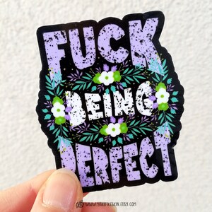 K&ouml;nnte beinhalten: Ein schwarzer und wei&szlig;er Aufkleber mit lila Schriftzug, der "F**k Being Perfect" lautet, umgeben von einem floralen Design mit gr&uuml;nen Bl&auml;ttern und wei&szlig;en Bl&uuml;ten.