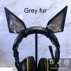 May include: Black gaming headset with grey fur cat ears attached. The headset has yellow accents and is on a stand. The text "Grey fur" is written above the headset. The text "Twitch, TikTok, Instagram" is written below the headset. The text "@missusyakut" is written below the social media icons.