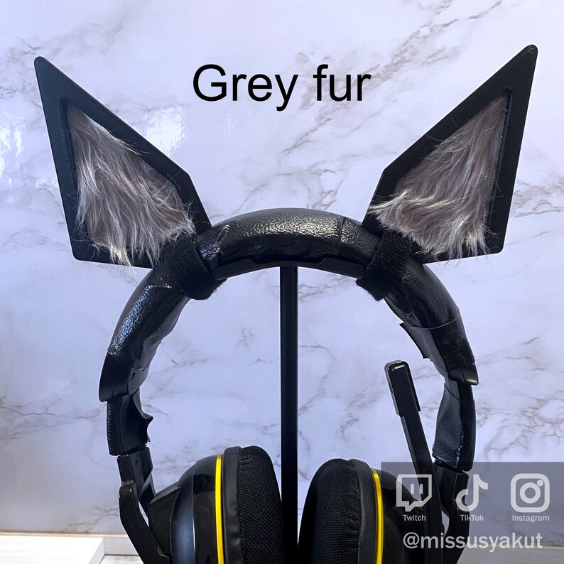 May include: Black gaming headset with grey fur cat ears attached. The headset has yellow accents and is on a stand. The text "Grey fur" is written above the headset. The text "Twitch, TikTok, Instagram" is written below the headset. The text "@missusyakut" is written below the social media icons.