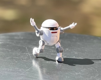 Puede incluir: Un juguete robot blanco con detalles grises y ojos rojos, de pie sobre una superficie gris con los brazos extendidos.