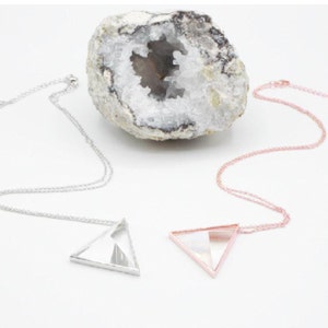 May include: Two silver necklaces with triangular pendants. The pendants are clear and have a geometric design. The necklaces are displayed on a white background with a rock in the background.