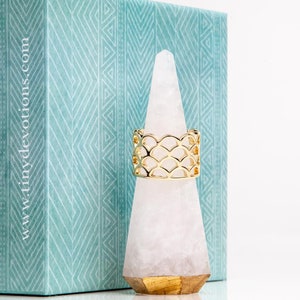 May include: Gold ring with a geometric design resembling fish scales. The ring is displayed on a white crystal ring holder.