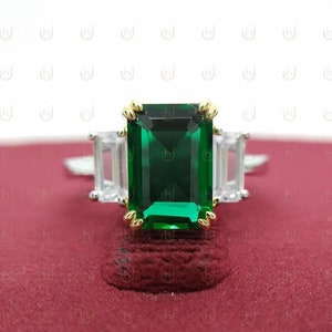 Pode incluir: Um anel de prata com uma grande pedra preciosa verde esmeralda de corte esmeralda, ladeada por duas pedras preciosas claras, retangulares e menores. A esmeralda é engastada em garras de ouro. O anel repousa sobre uma superfície vermelha.