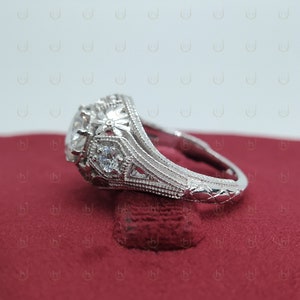 May include: A silver engagement ring with a large, round center stone and smaller accent stones. The ring has a detailed, intricate design with floral motifs.