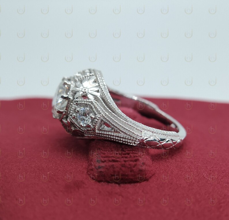May include: A silver engagement ring with a large, round center stone and smaller accent stones. The ring has a detailed, intricate design with floral motifs.