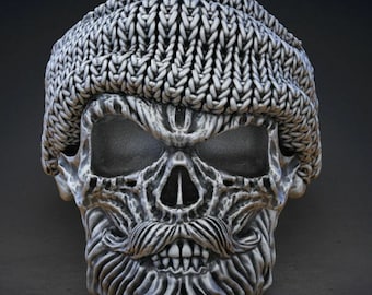 Op de afbeelding: Een zilveren schedelring met een gebreide beanie op de bovenkant en een gedetailleerde baard en snor. De schedel heeft een dreigende uitdrukking met donkere oogkassen.