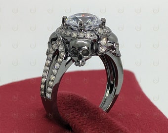 May include: Black skull ring with a large clear cubic zirconia stone. The ring is set in black metal and has a skull design on the band.