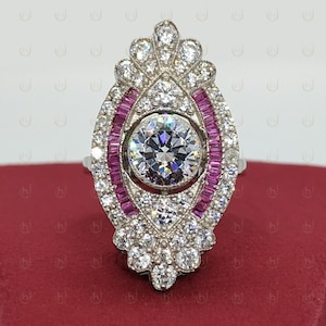 May include: A silver ring with a large round center stone surrounded by smaller stones. The ring has a decorative design with pink stones around the center stone.