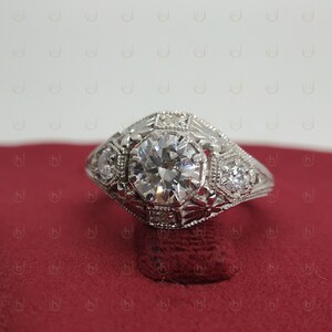 May include: A silver engagement ring with a large, round, clear diamond in the center. The ring has a detailed, intricate design with smaller diamonds surrounding the center stone.