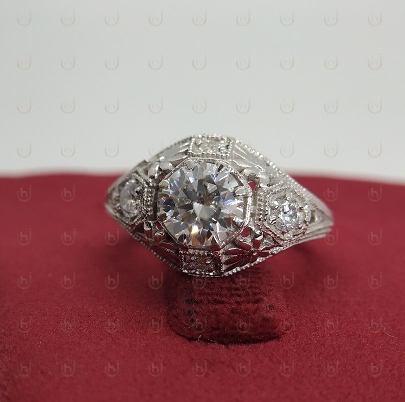 May include: A silver engagement ring with a large, round, clear diamond in the center. The ring has a detailed, intricate design with smaller diamonds surrounding the center stone.