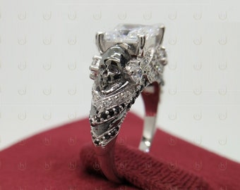 May include: A silver ring with a black skull design and a large clear cubic zirconia stone. The ring is set on a red velvet background.