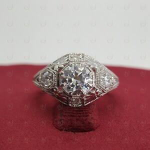 May include: A silver engagement ring with a large, round, clear diamond center stone. The ring has a detailed, intricate design with smaller diamonds surrounding the center stone.