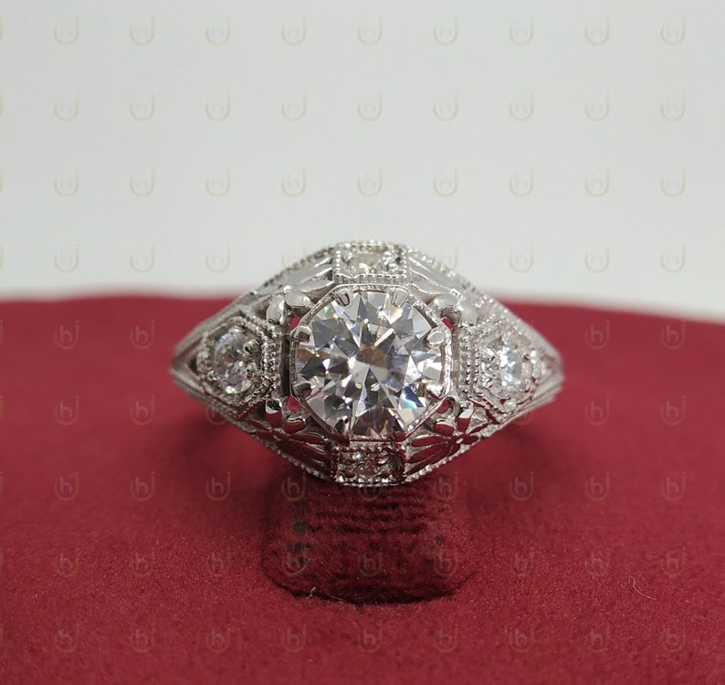 May include: A silver engagement ring with a large, round, clear diamond center stone. The ring has a detailed, intricate design with smaller diamonds surrounding the center stone.