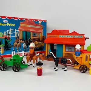 Op de afbeelding: Vintage Fisher-Price Western Town speelset met een doos, gebouwen, figuren, paarden en wagens. De set bevat een rood en oranje Western Town gebouw, een groene wagen en een gele postkoets. De doos is blauw met het Fisher-Price logo.