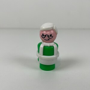 Op de afbeelding: Een vintage Fisher-Price Little People figuur van een vrouw. De figuur heeft een roze gezicht met een zwarte bril en een witte hoed. Het lichaam is groen met een wit harnas.