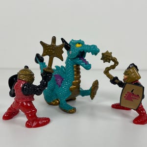 Op de afbeelding: Een talingkleurig drakenfiguur met gouden accenten en paarse vleugels staat tussen twee kleine krijgersfiguren. De krijgers dragen rode harnassen en gouden helmen, en houden elk een bruin wapen en een schild vast, met een drakenafbeelding.