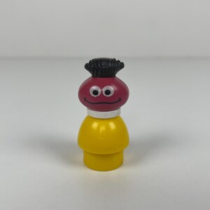 Pode incluir: Uma figura vintage Fisher-Price Little People. A figura tem um corpo amarelo, uma cabeça vermelha com cabelo preto, um pescoço branco e um rosto sorridente. Um brinquedo colecionável clássico.
