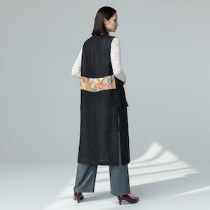 May include: A long black vest with a colorful floral design across the back. The vest is worn over a white lace-sleeved top and gray pants. The vest has a side pocket and a slit on the side.