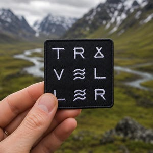 Op de afbeelding: Een zwart vierkant embleem met witte geborduurde tekst 'TRAVELER' in een gestileerd ontwerp. De patch wordt vastgehouden voor een schilderachtig berglandschap met een rivier en een bewolkte lucht.