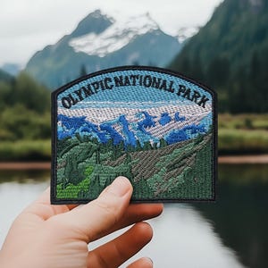 May include: Embroidered patch featuring the text "OLYMPIC NATIONAL PARK" above a scenic mountain landscape. The patch has a black border, blue sky, and green and blue mountain imagery. The patch is held up in front of a lake and mountain backdrop.