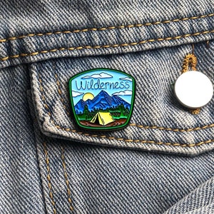 Op de afbeelding: Een blauwe en groene emaille pin met een berglandschap en het woord "Wilderness" in wit schrift. De pin is op een blauwe denim jas.