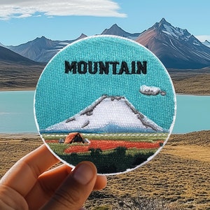 Op de afbeelding: Een blauwe en witte geborduurde patch met het woord "MOUNTAIN" in zwarte letters. De patch toont een witte berg met een kleine oranje tent aan de voet. De patch wordt vastgehouden voor een schilderachtig landschap met een meer en bergen op de achtergrond.