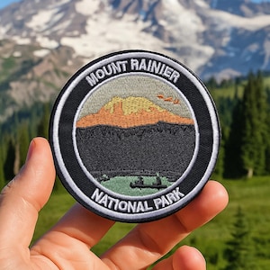 Può includere: Una toppa rotonda con la scritta "Mount Rainier National Park". La toppa presenta un'illustrazione di una catena montuosa, un lago e alberi. La toppa è nera con un bordo bianco. È tenuta in mano.