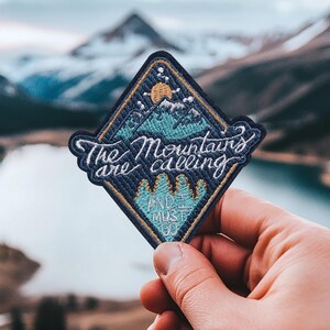 Op de afbeelding: Een blauwe, ruitvormige geborduurde patch met de tekst "The Mountains are calling and I must go". De patch toont een bergketen met een zon en sterren.