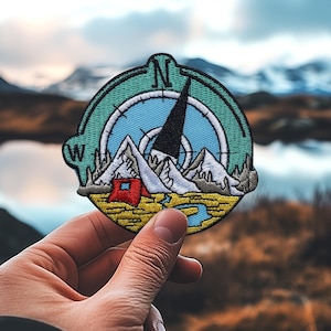 Könnte beinhalten: Ein blauer und gelber Kompass-Patch mit einem roten Zelt und Bergen, die darauf gestickt sind. Der Kompass hat eine schwarze Nadel, die nach Norden zeigt, und die Buchstaben N, W und S sind sichtbar.