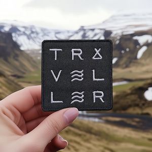 Könnte beinhalten: Schwarzer Patch mit weiß gesticktem Text, der "TR&VL LR" lautet, mit einem Wellensymbol zwischen dem "V" und dem "L" und einem Dreiecksymbol zwischen dem "R" und dem "V".