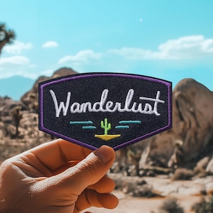 May include: A purple and black embroidered patch with the word "Wanderlust" in white lettering. A small green cactus is embroidered in the centre of the patch.