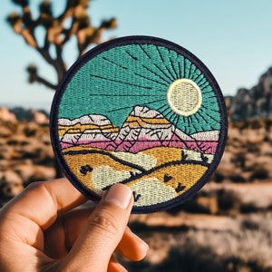 Op de afbeelding: Een ronde geborduurde patch met een woestijnlandschap. De patch toont een bergketen, een zon en een blauwe lucht met lichtstralen. De bergen zijn roze, geel en wit, en de lucht is een levendige turquoise.