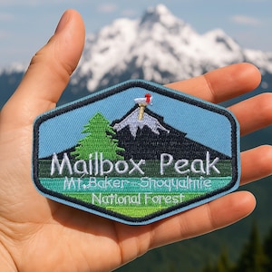 May include: Embroidered patch featuring a mountain scene with the text "Mailbox Peak" and "Mt. Baker - Snoqualmie National Forest." The patch has a blue background with a mountain, tree, and mailbox design. The patch is held in a hand.