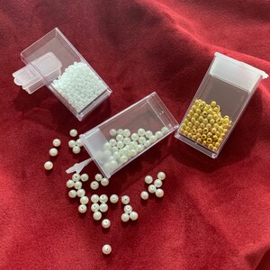 May include: Three clear plastic containers filled with white, gold, and white beads. The containers are on a red surface. Some white beads are scattered on the surface.