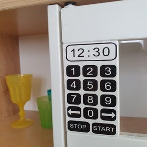 Könnte beinhalten: Weißer Spielzeug-Mikrowellenherd mit einem schwarzen digitalen Display, das 12:30 anzeigt. Die Mikrowelle hat eine Zifferntastatur mit Tasten für die Zahlen 1 bis 9, 0 und Pfeiltasten zum Starten und Stoppen. Die Mikrowelle hat auch eine Stopp- und Starttaste.