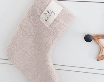 May include: A beige sherpa fleece Christmas stocking featuring a rectangular fabric tag sewn onto the upper section. The tag displays the name Lily in a black cursive script font. The stocking is hung from a black circular knob.