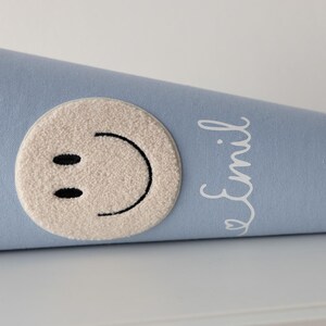 May include: A light blue cone-shaped gift bag featuring a cream-coloured tufted circular patch with a black embroidered smiley face. The name Emil is printed in white cursive script on the side of the fabric cone, which has a cream fabric trim.