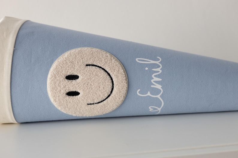 May include: A light blue cone-shaped gift bag featuring a cream-coloured tufted circular patch with a black embroidered smiley face. The name Emil is printed in white cursive script on the side of the fabric cone, which has a cream fabric trim.