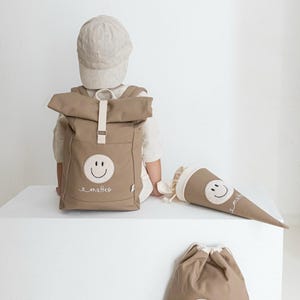 May include: A tan roll-top backpack, a cone-shaped gift bag, and a drawstring bag, all featuring a white circular patch with a black embroidered smiley face and the name "matteo" in white cursive script, paired with a tan baseball cap.
