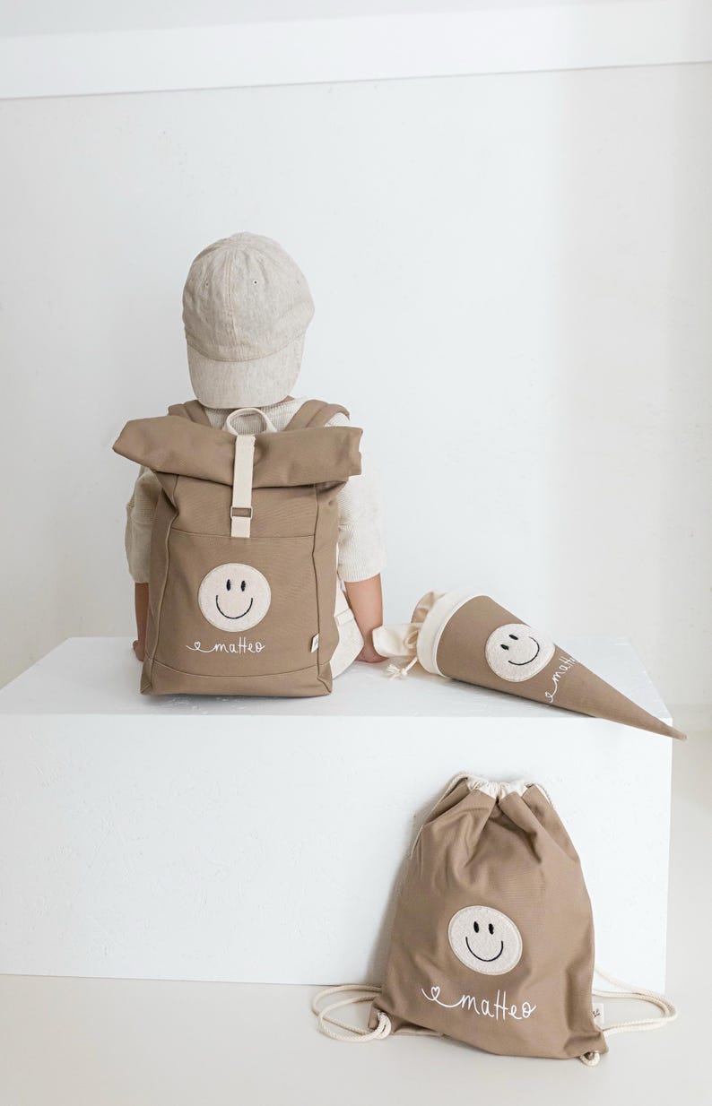 May include: A tan roll-top backpack, a cone-shaped gift bag, and a drawstring bag, all featuring a white circular patch with a black embroidered smiley face and the name "matteo" in white cursive script, paired with a tan baseball cap.