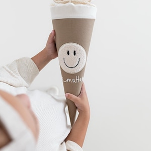 May include: A beige cone-shaped gift bag with a cream-coloured fabric top secured by a drawstring. The bag features a circular white textured patch with a black embroidered smiley face and the word "matter" written in white cursive script below it.