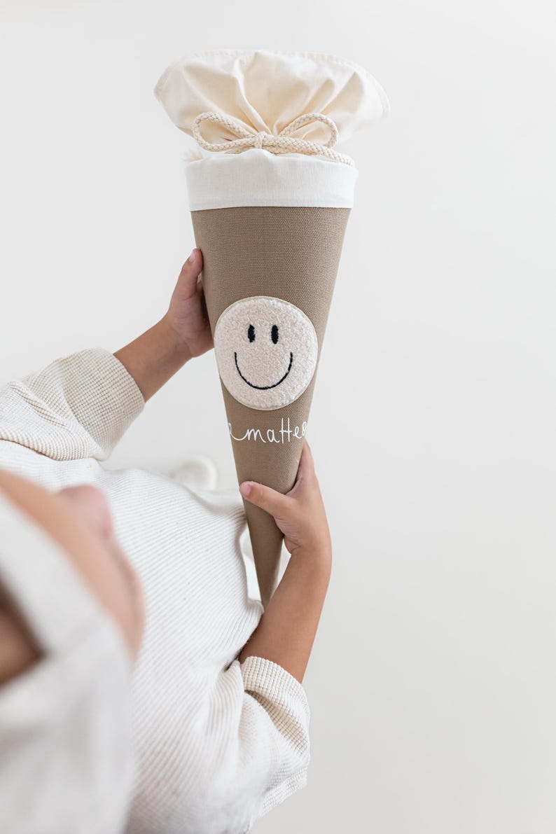 May include: A beige cone-shaped gift bag with a cream-coloured fabric top secured by a drawstring. The bag features a circular white textured patch with a black embroidered smiley face and the word "matter" written in white cursive script below it.