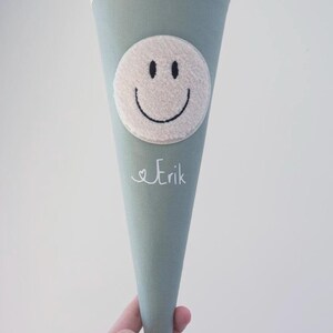 May include: A sage green cone-shaped fabric gift bag featuring a cream-coloured plush circular smiley face patch and the name Erik embroidered in white script. The top of the bag is finished with a cream-coloured fabric cuff and drawstring closure.