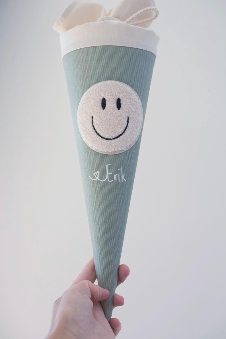 May include: A sage green cone-shaped fabric gift bag featuring a cream-coloured plush circular smiley face patch and the name Erik embroidered in white script. The top of the bag is finished with a cream-coloured fabric cuff and drawstring closure.