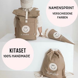 Könnte beinhalten: Ein beigefarbener Rolltop-Rucksack, eine Schultüte und ein Turnbeutel, jeweils mit einem weißen runden Aufnäher mit schwarzem Smiley-Gesicht und dem Namen matteo in weißer Schreibschrift. Text: NAMENSPRINT VERSCHIEDENE FARBEN und KITASET 100% HANDMADE.