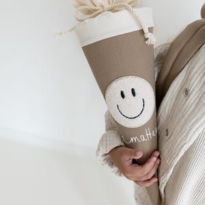 May include: A beige cone-shaped fabric gift bag featuring a white circular patch with a black embroidered smiley face. The top of the bag is made of off-white fabric with a drawstring closure. The word matter is embroidered in white cursive script.