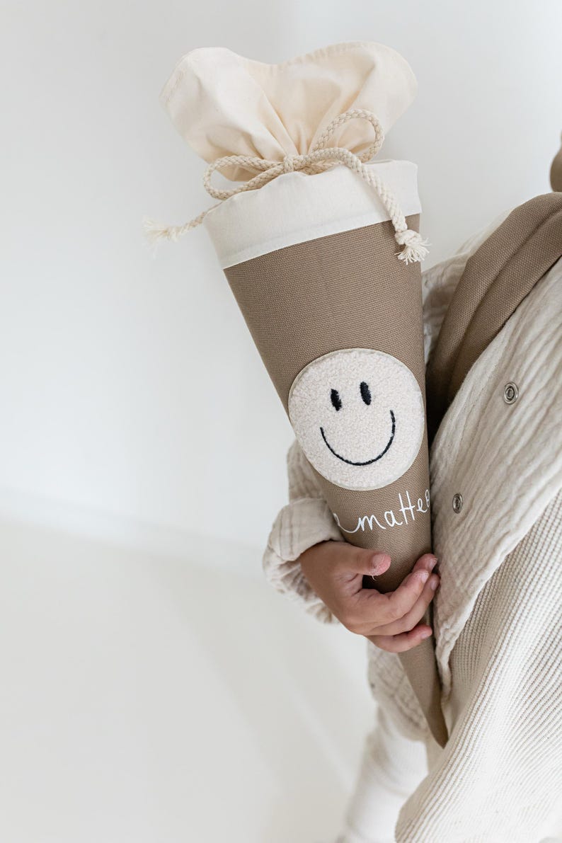 May include: A beige cone-shaped fabric gift bag featuring a white circular patch with a black embroidered smiley face. The top of the bag is made of off-white fabric with a drawstring closure. The word matter is embroidered in white cursive script.
